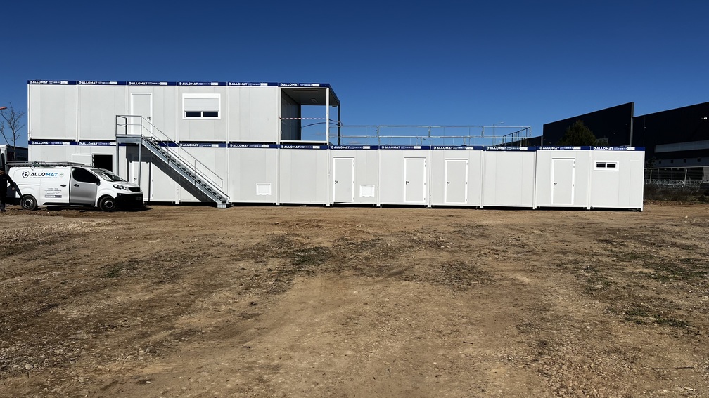 Une base vie de chantier composée de 18 bungalows dont 6 en R+1 avec une terrasse et garde-corps. Une camionnette Allomat est garée devant.