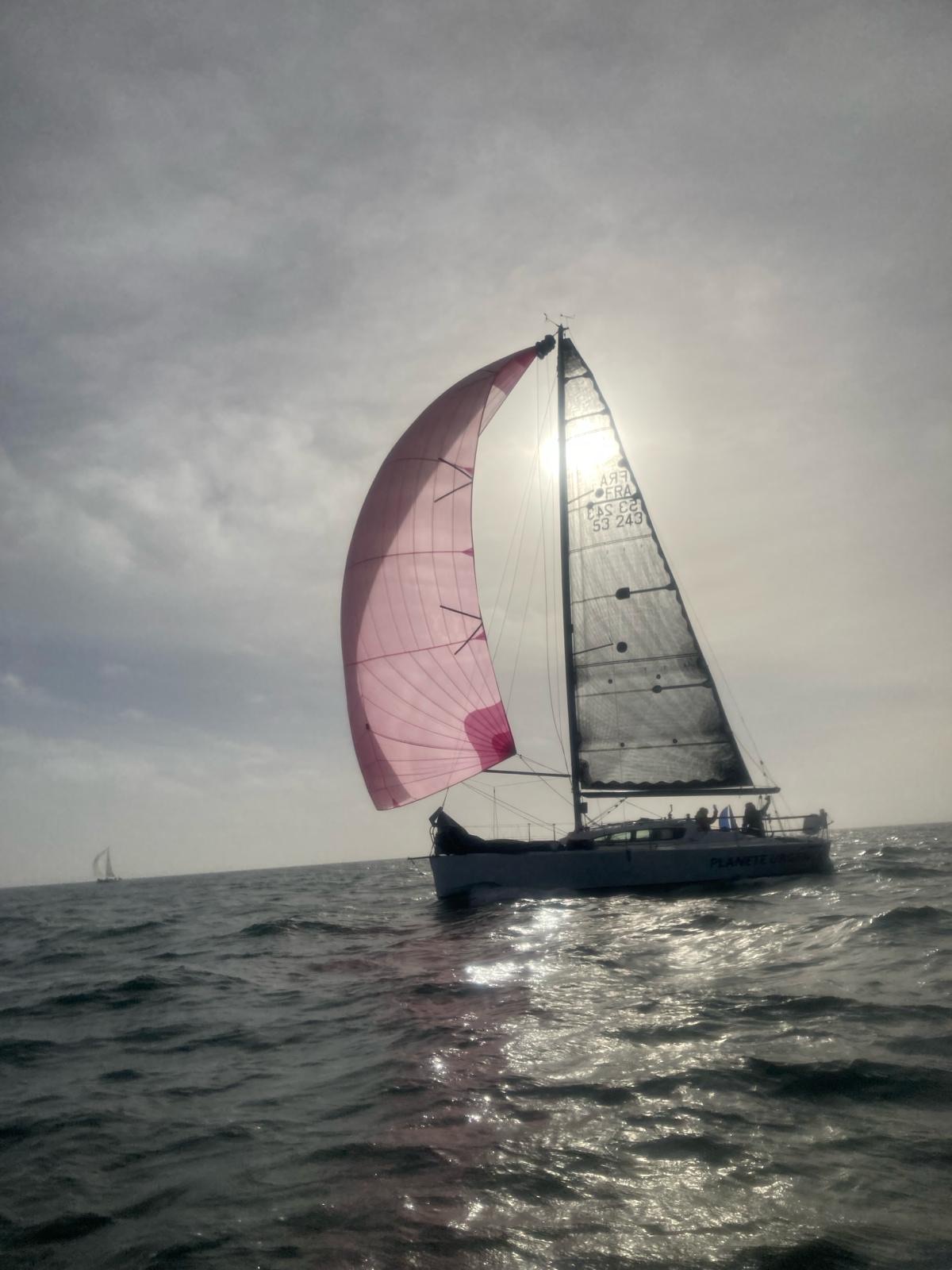 Un voilier sur la mer, en contre-jour. Le soleil filtre à travers la voile de droite, blanche. Celle de gauche est rouge