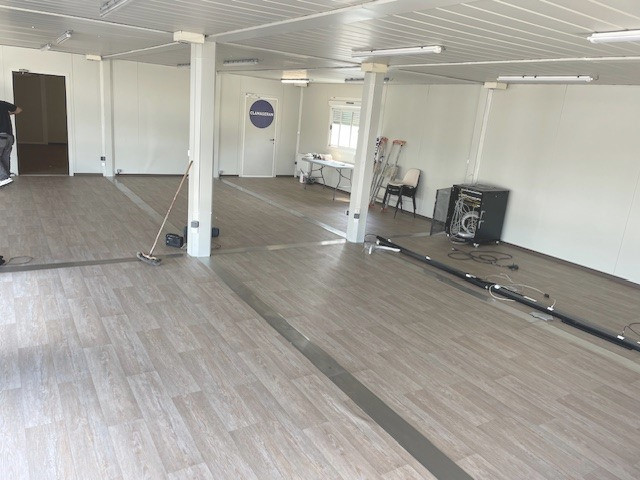 Des bureaux modulaires vides avec un sol PVC façon parquet et des poteaux au milieu de la grande pièce, qui sont en fait les montants des modules temporaires. Les jointures entre modules sont fait avec de belles finitions métallisées. Les murs au fond la pièce sont gris clairs