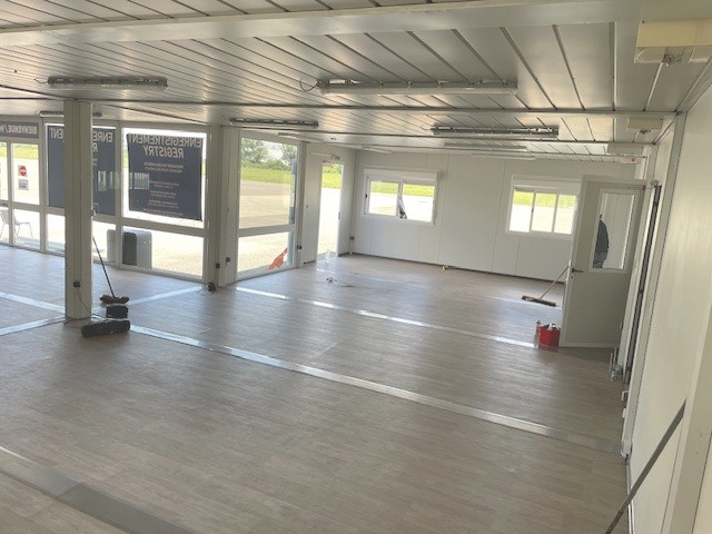 Des bureaux modulaires vides avec un sol PVC façon parquet et des poteaux au milieu de la grande pièce, qui sont en fait les montants des modules temporaires. Les modules sont repeints en gris. Les jointures entre modules sont fait avec de belles finitions métallisées. On peut voir une belle baie vitrée avec un covering au nom de Eurexpo Lyon